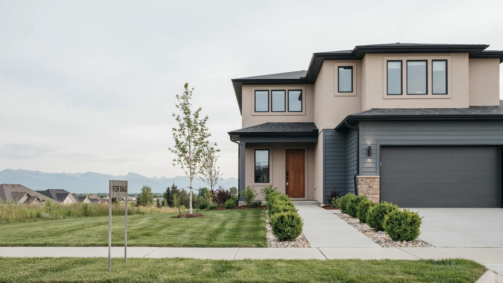 A modern two storey home with mountains behind it and a for sale sign on the lawn, the Lethbridge foreclosure guide